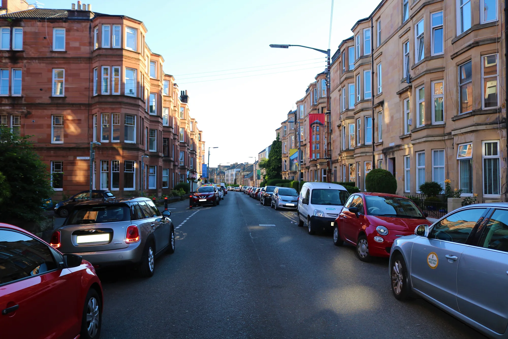 A street of red and blond sandstone tenements with bay windows on Deanston Drive, Shawlands, Glasgow, with cars parked along both sides