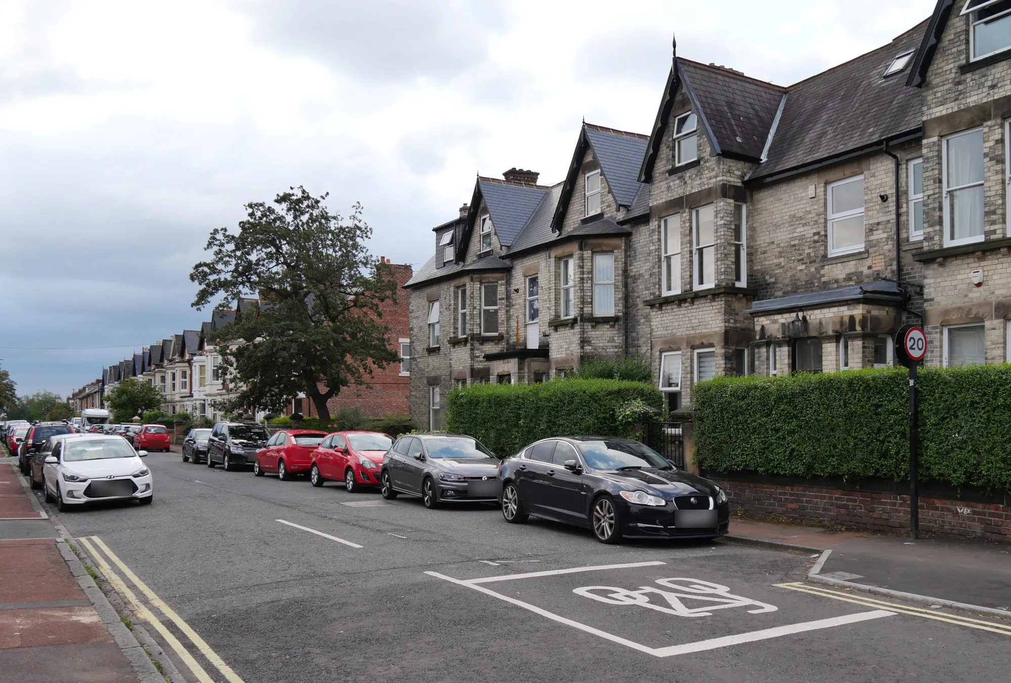A row of stone-fronted terraced houses with bay windows and hedges on Rothbury Terrace, Heaton, Newcastle upon Tyne, with cars parked along the street