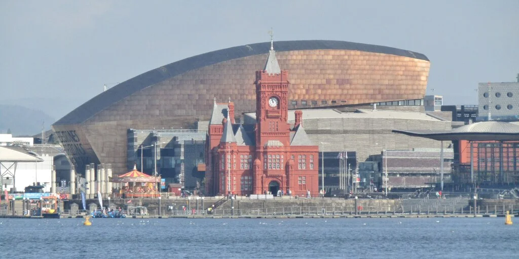 The red-brick Pierhead Building with its clock tower in front of the curved, copper-coloured Wales Millennium Centre, seen across the water of Cardiff Bay
