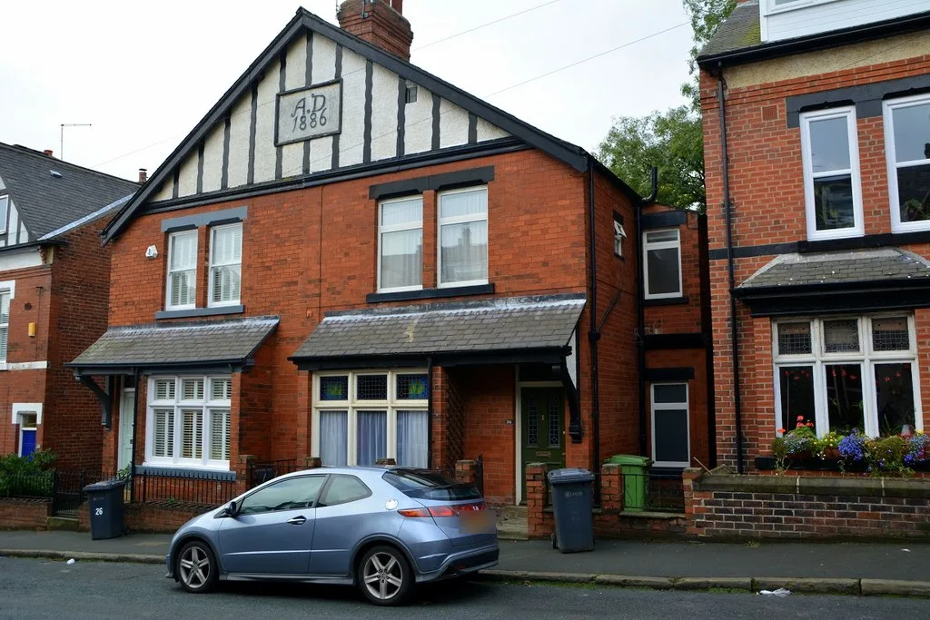 Red-brick semi-detached houses on Northbrook Street, Chapel Allerton, with a black-and-white timbered gable dated A.D. 1886, slate porch canopies, wheelie bins and a parked silver hatchback