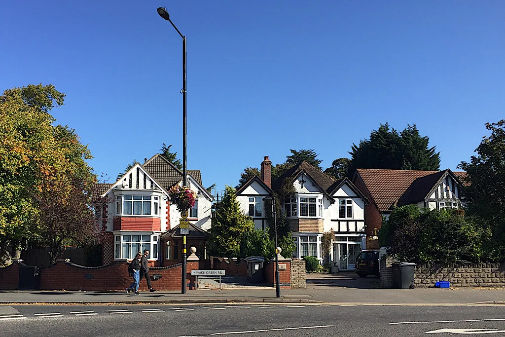 Detached houses with white render, bay windows and tiled roofs behind low brick walls on Wake Green Road, Moseley, with a Wake Green Rd street sign and a street lamp in front